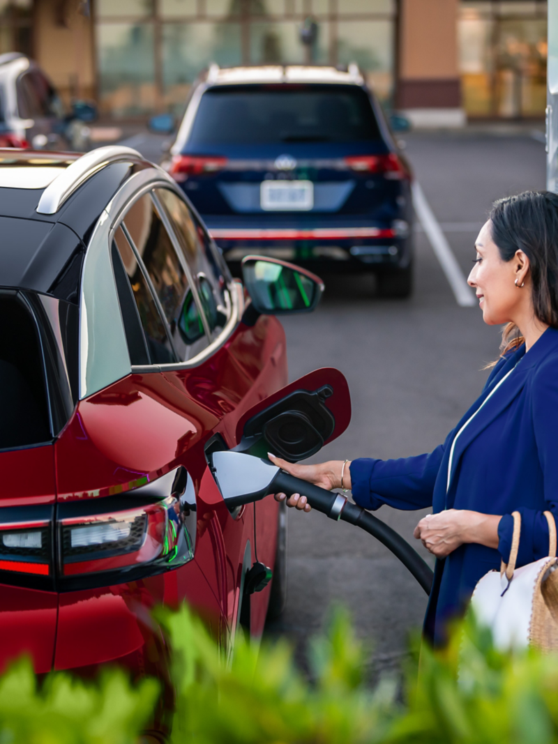 A well-dressed woman, parked at charging station stands on the passenger side of an ID.4 SUV in Aurora Red Metallic plugging a charging cable into the vehicle.