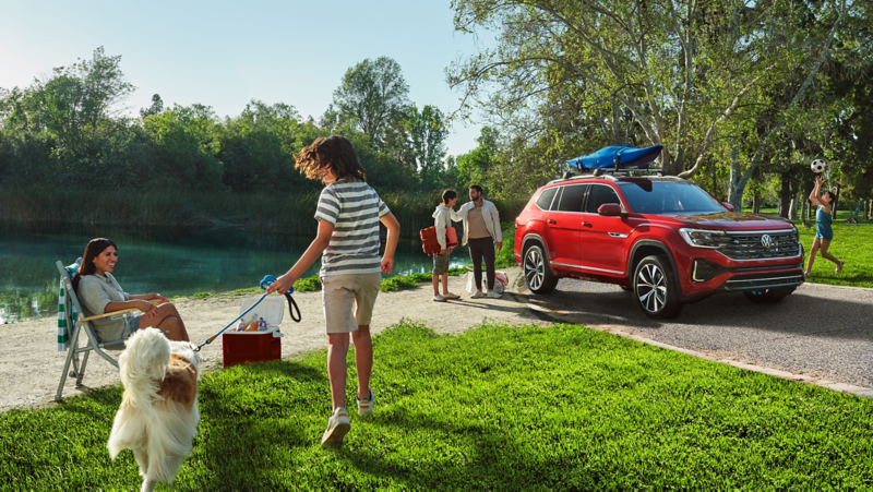 Atlas se muestra en Aurora Red Metallic estacionado en la acera en un entorno de parque con la familia disfrutando del parque y el lago de fondo.