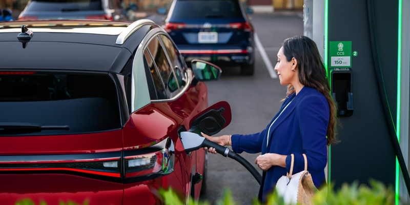 An ID.4 in Aurora Red Metallic as seend from the rear with a woman connecting the charging device.