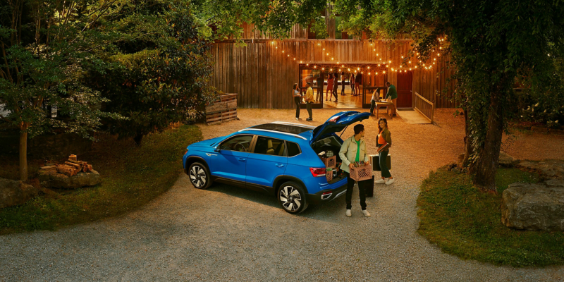 A couple unpacks a crate of vinyl records and DJ equipment from the back of a Taos in Cornflower Blue that’s parked on a driveway stringed with lights.