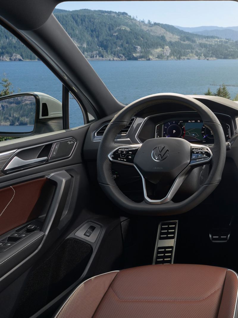 A view of the Tiguan interior in Noisette Brown Vienna leather seating surfaces, as seen from the driver seat with mountains and a lake in the background.
