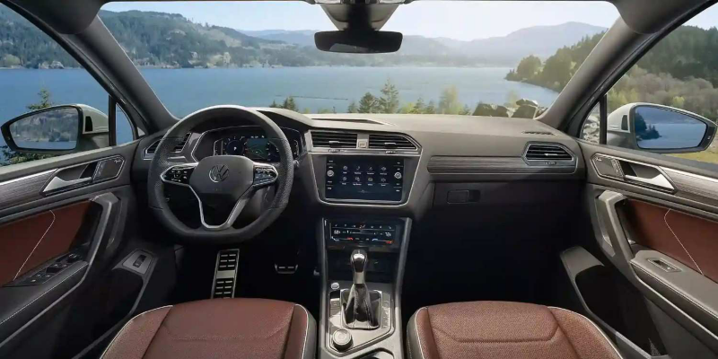 A view of the Tiguan interior in Noisette Brown Vienna leather seating surfaces, as seen from the driver seat with mountains and a lake in the background.