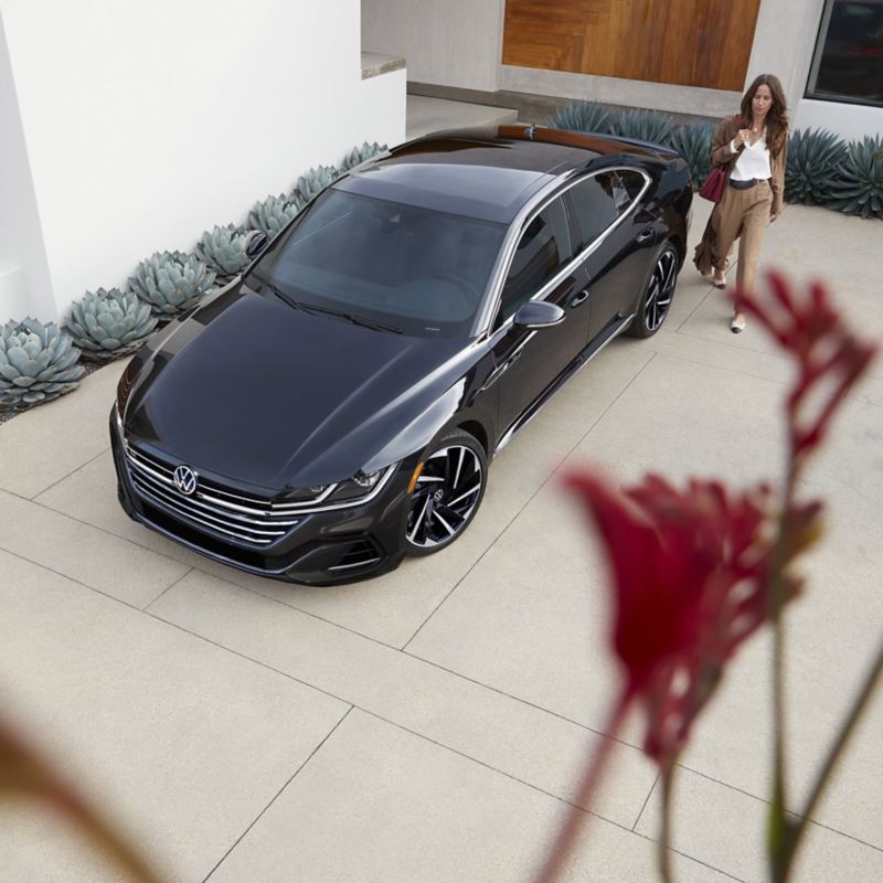 Arteon shown in Deep Black Pearl parked in the driveway of a modern home with woman walking to Arteon