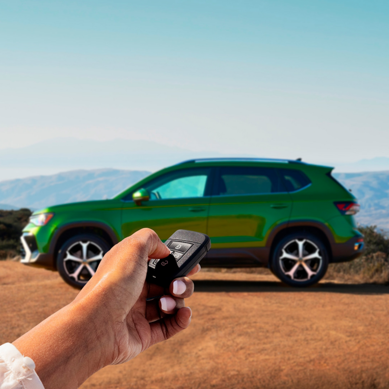 A hand holding a VW key fob with a Taos shown in Bright Moss Green parked in the background.