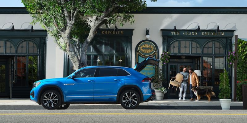 A side profile view of the Atlas Cross Sport shown in Kingfisher Blue Metallic parked in front of an antique shop with the rear hatch open and a couple and their dog walking towards with one carrying a chair.