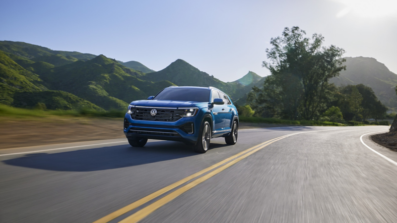 A front ¾ view of an Atlas Cross Sport shown in Kingfisher Blue Metallic driving on a road with trees and mountains in the background.