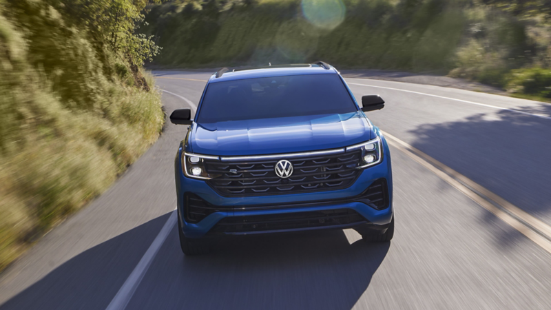 A front view of an Atlas Cross Sport shown in Kingfisher Blue Metallic driving on a road with trees and mountains in the background.