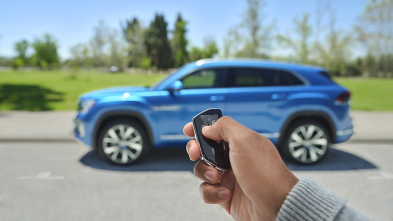 A close-up view of a person holding a key fob with the Atlas Cross Sport shown in Kingfisher Blue Metallic parked next to a grassy knoll and trees in the background.
