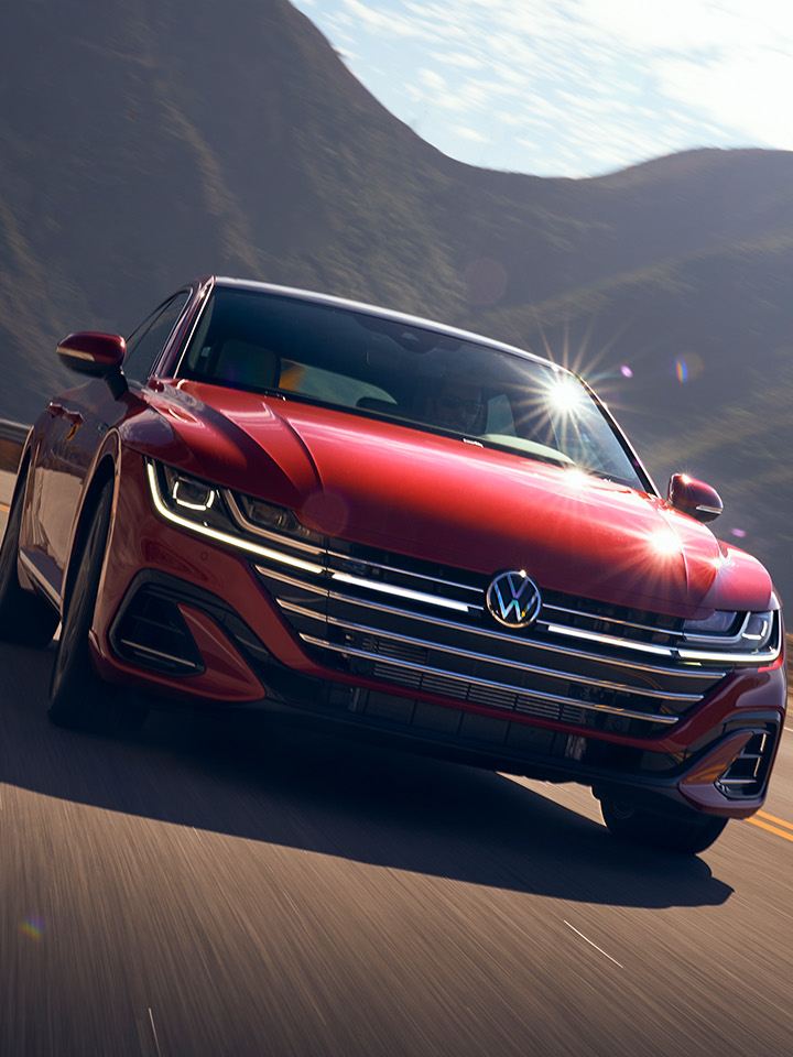 A front view of an Arteon shown in Kings Red Metallic, driving on a two-lane road with mountains behind it. 