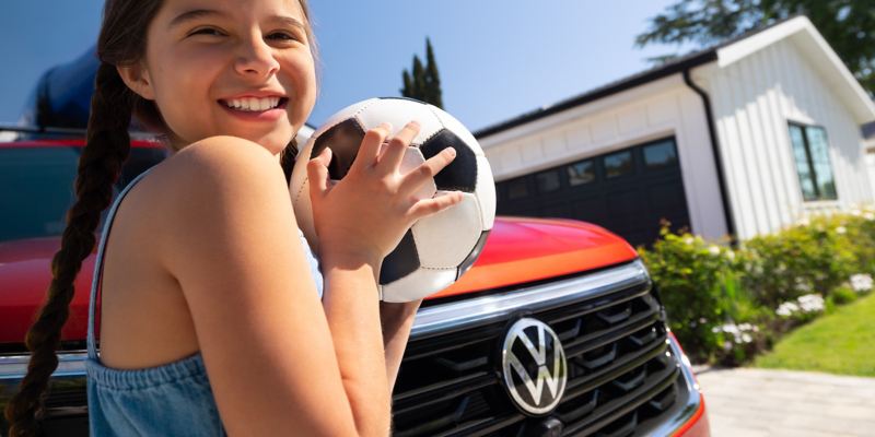 Primer plano de una joven sonriendo y sujetando un balón de fútbol con un Atlas estacionado en la entrada de una casa al fondo.