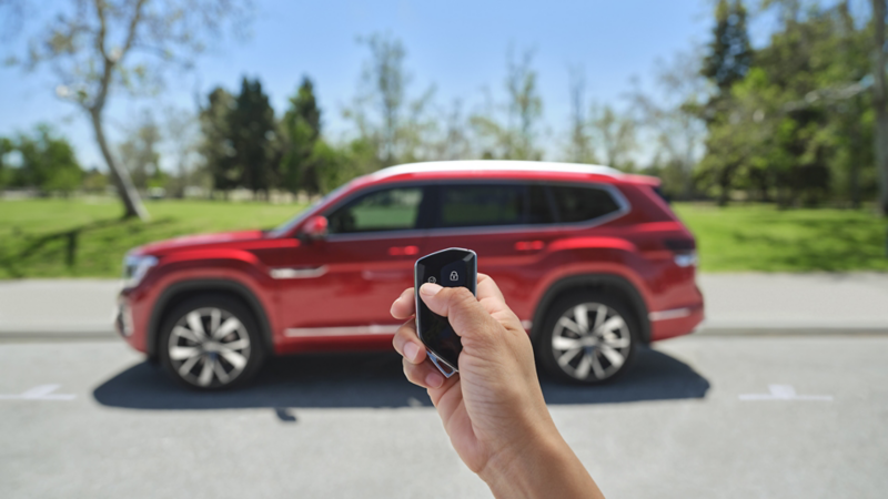 A hand holding a VW key fob with an Atlas shown in Aurora Red Metallic parked in the background.