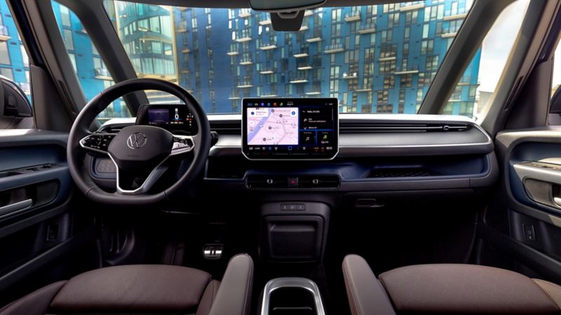 View from the front seat of an ID. Buzz in Moonlight interior showing the front seats, steering wheel, infotainment display and dash with city lights visible through the windshield.