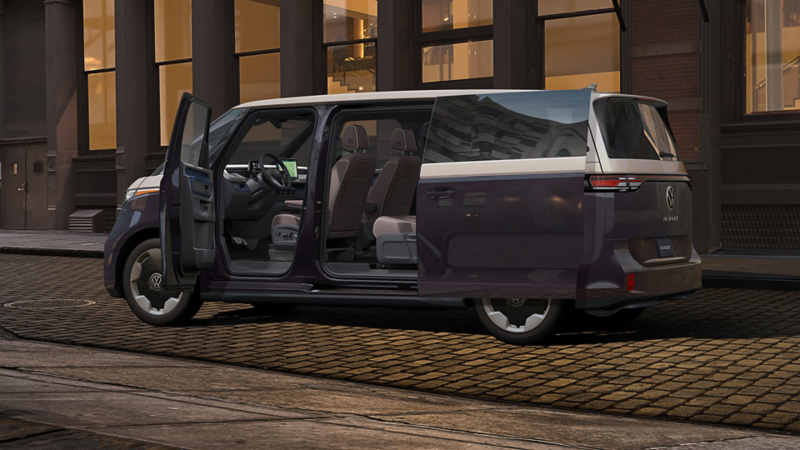 An ID. Buzz shown in Blue Charcoal Metallic and Candy White with the front and side sliding doors open is parked on a city street in the evening.