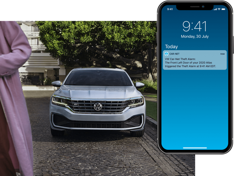 Exterior front view of a Passat in Reflex Silver Metallic parked next to a lawn with driver and passenger walking away. On their mobile phone, we see a Volkswagen Car-Net® Anti-Theft Alert.