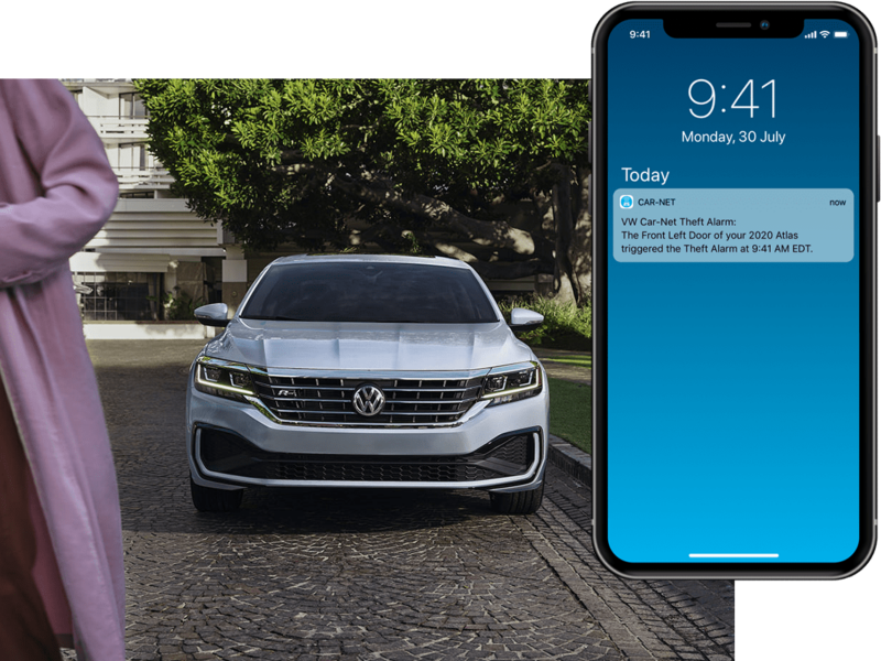 Exterior front view of a Passat in Reflex Silver Metallic parked next to a lawn with driver and passenger walking away. On their mobile phone, we see a Volkswagen Car-Net® Anti-Theft Alert.