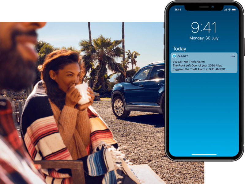 A man and women enjoy a mug of coffee outside their VW. On their mobile phone, we see a Volkswagen Car-Net® Safe & Secure alert.