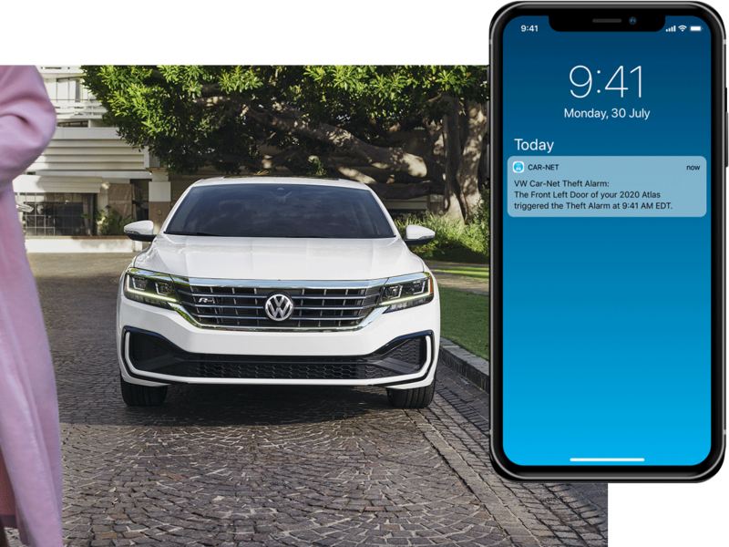 Exterior front view of a Passat in Pure White parked next to a lawn with driver and passenger walking away. On their mobile phone, we see a Volkswagen Car-Net® Anti-Theft Alert.