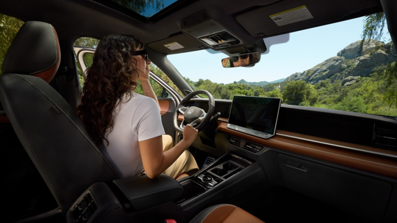 Interior view from the front passenger side of a parked Tiguan shown in Amber Brown leather with a woman exiting the driver's seat and a lush forest and rock outcroppings visible through the windshield.
