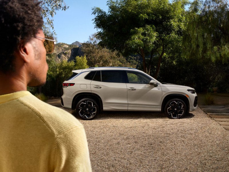 View over the shoulder of a man with sunglasses looking at a profile angle of a Tiguan shown in Pure White parked with trees in the background. 