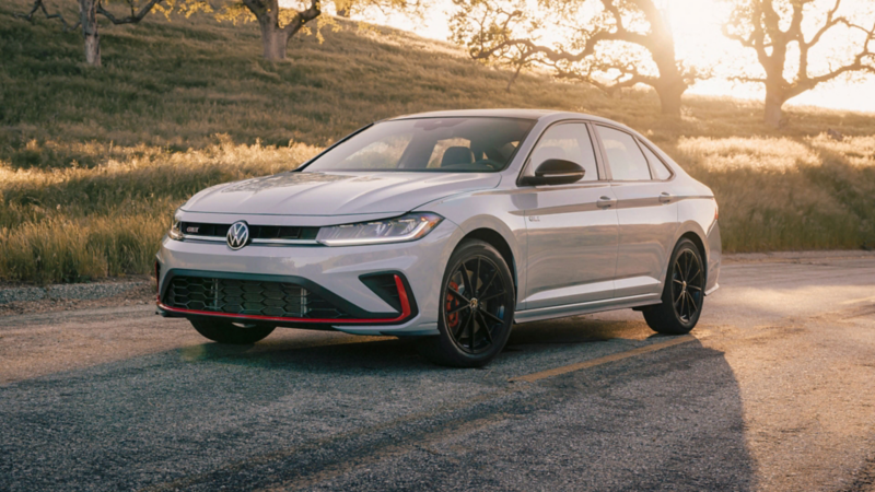 Vista de 3/4 frontal de un Jetta GLI en color Monument Gray estacionado frente a una colina verde con árboles al fondo.