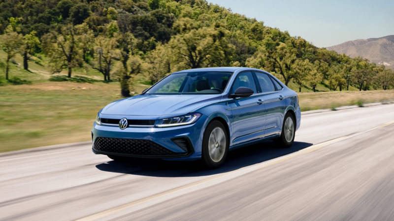 Front ¾ view of a Jetta shown in Monterey Blue Pearl driving on an open road with wooded hills in the background.