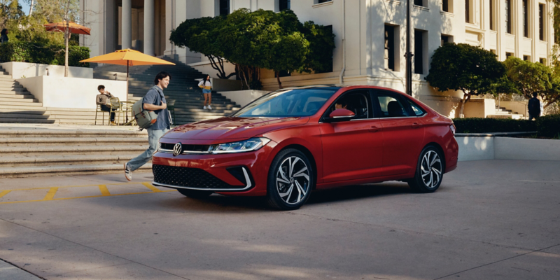 A Jetta shown in Kings Red Metallic is parked at a college campus while a student walks toward the Jetta.