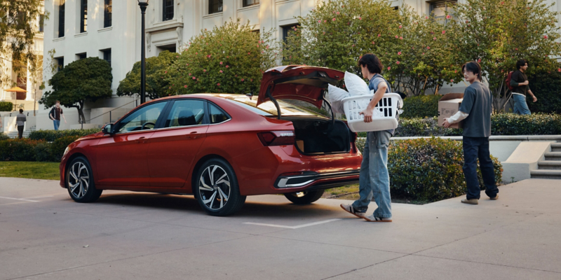 A Jetta shown in Kings Red Metallic is parked at a college campus while two students unload personal items from the trunk.