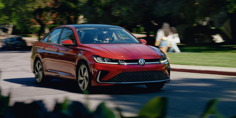 Front view of a Jetta shown in Kings Red Metallic driving through a college campus with cars, trees and people walking in the background