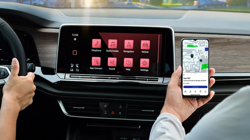 The front passenger in a parked Volkswagen holds a smartphone in front of the center touchscreen display.