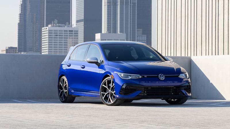 A Lapiz Blue Metallic Golf R is parked on the roof-level of a downtown parking garage. A city skyline with tall buildings can be seen in the distance.