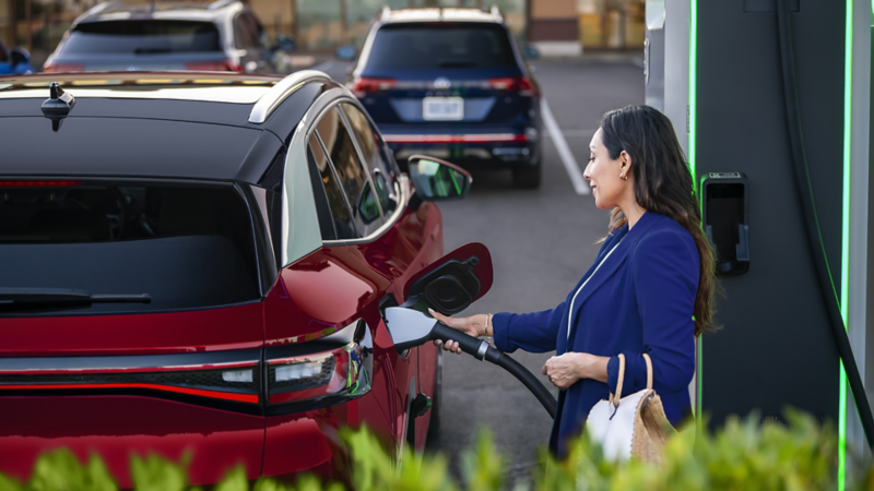 A well-dressed woman, parked at charging station stands on the passenger side of an ID.4 a SUV in Aurora Red Metallic plugging a charging cable into the vehicle.