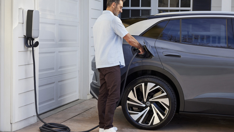 Rear side view of an ID.4 in Pure Gray parked in front of a home garage with a man plugging in the home charger.