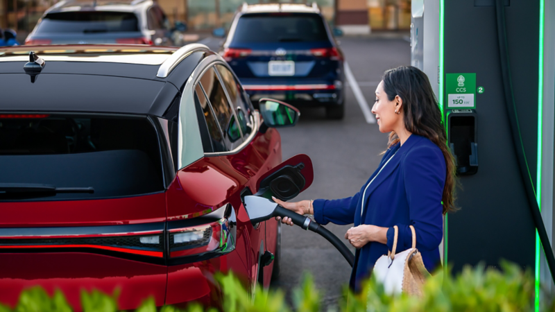 A well-dressed woman, parked at charging station stands on the passenger side of an ID.4 SUV in Aurora Red Metallic plugging a charging cable into the vehicle.