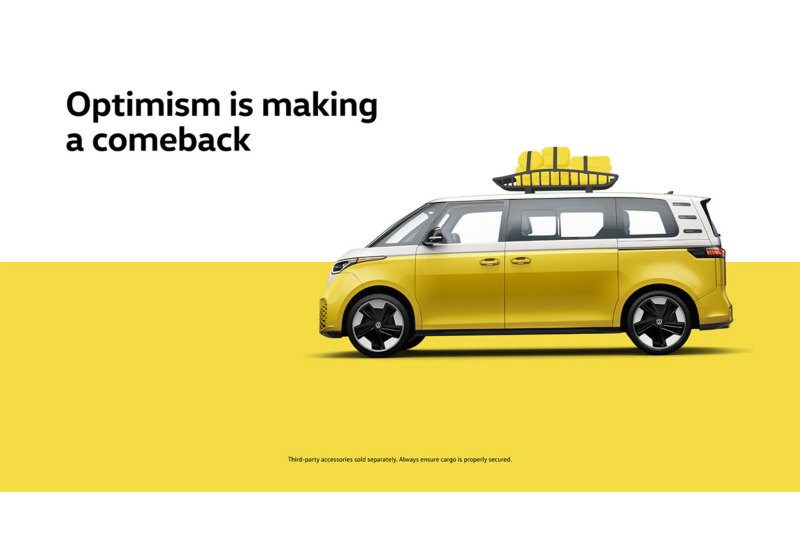 A side view of the ID. Buzz in Pomelo Yellow Metallic with a luggage rack and luggage strapped in it over a two-tone white and yellow background with the words “Optimism is making a comeback” to the left of the vehicle. 