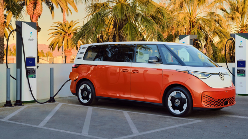 Side view of an ID. Buzz shown in Energetic Orange Metallic and Candy White parked at a charging station with palm trees in the background.