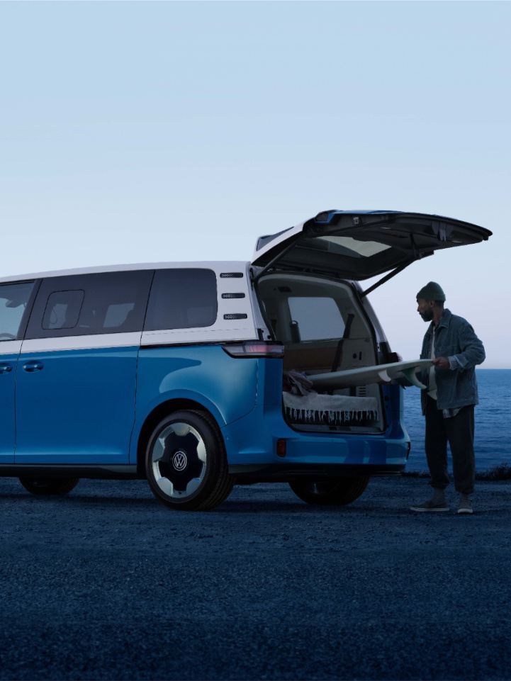 An ID. Buzz shown in Cabana Blue Metallic at a beach with surfers outside the vehicle and the ocean in the background.