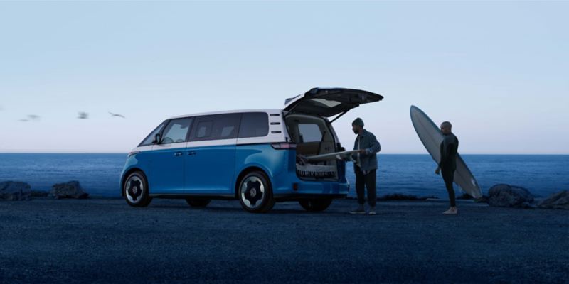 An ID. Buzz shown in Cabana Blue Metallic at a beach with surfers outside the vehicle and the ocean in the background.