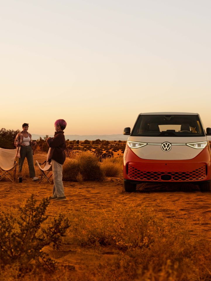 An ID. Buzz shown in Energetic Orange Metallic in a desert landscape with people outside the vehicle preparing to enjoy the outdoors.