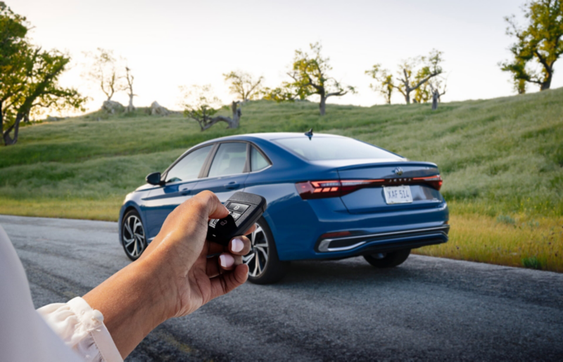 Rear view of a Jetta shown in Monterey Blue Pearl parked with trees in the background and a person holding a VW key fob in the foreground.