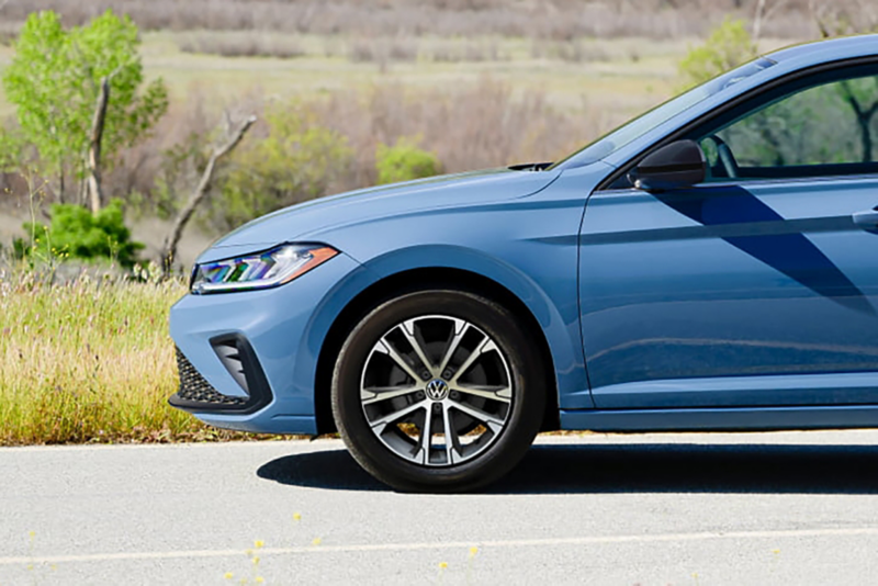 Profile view of a parked Jetta shown in Monterey Blue Pearl highlighting the driver side front wheel with trees in the background. 