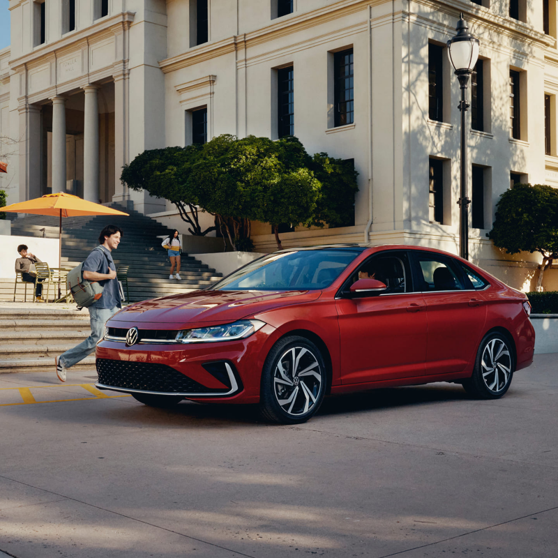 A Jetta shown in Kings Red Metallic is parked at a college campus while a student walks toward the Jetta.