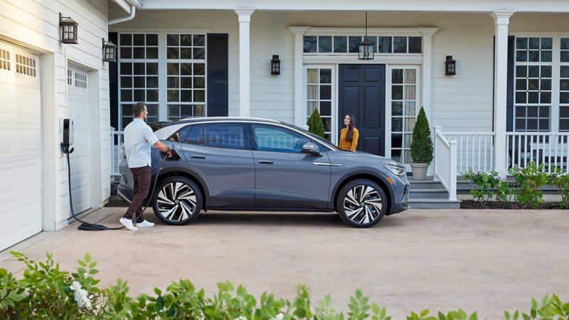 Side profile view of an ID.4 in Pure Gray parked in front of a home with a man plugging in the home charger and a woman standing at the front door. 