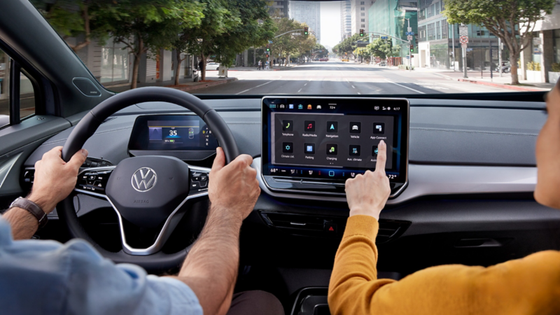 A close-up shot of an ID.4 dashboard over the shoulders of the driver and passenger, focusing on the center display as the passenger reaches out to the touchscreen.
