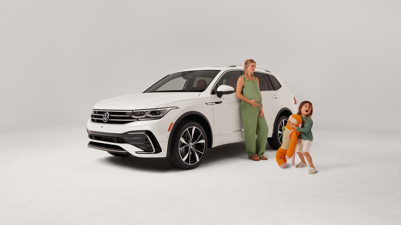 Front 3/4 view of a Tiguan in Opal White parked on a white background with a mom leaning on the driver door and a kid holding a stuffed animal.