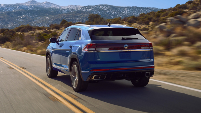 Rear view of an Atlas Cross Sport shown in Mountain Lake Blue driving on a road with mountains in the background.