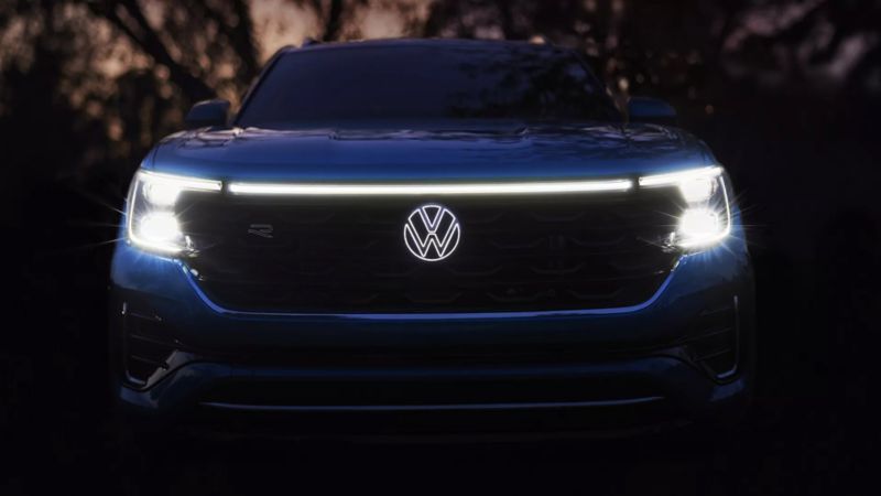 A close-up front view of the automatic LED headlights and available illuminated VW emblem on an Atlas Cross Sport shown in Mountain Lake Blue at sunset.