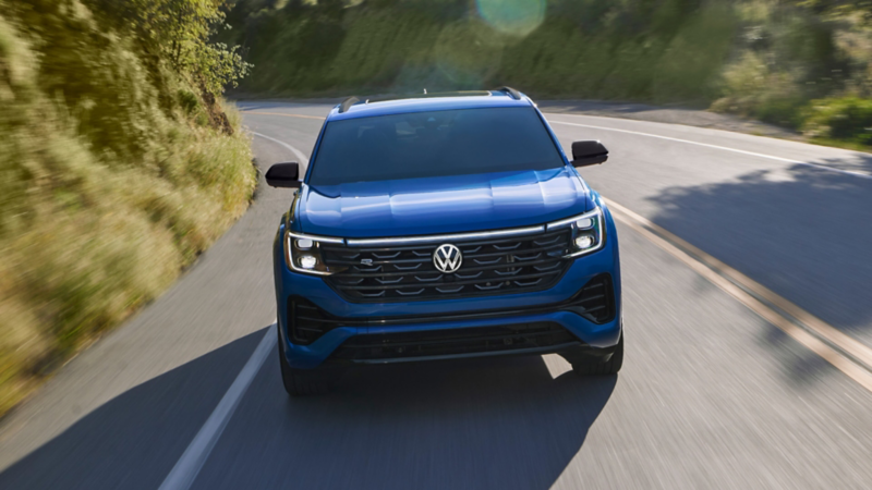 Vista frontal de un Atlas Cross Sport en color Mountain Lake Blue Metallic circulando por una carretera boscosa de montaña.