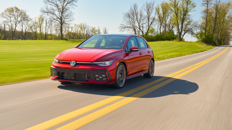 Front view of a Golf GTI shown in Kings Red Metallic driving on a country highway next to green grass with trees in the distance.