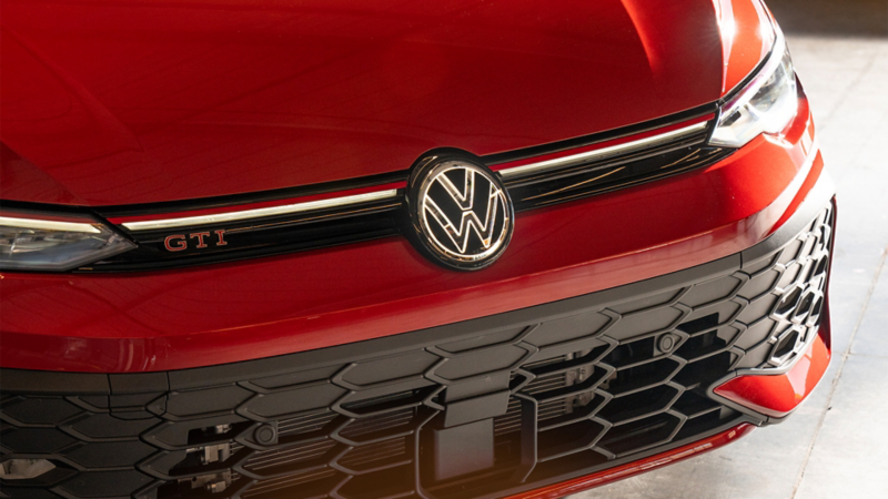 Close-up of the front passenger's side of a Kings Red Metallic Golf GTI Autobahn parket in a concrete floor, highlighting the LED headlight, honeycomb grille, and VW logo.