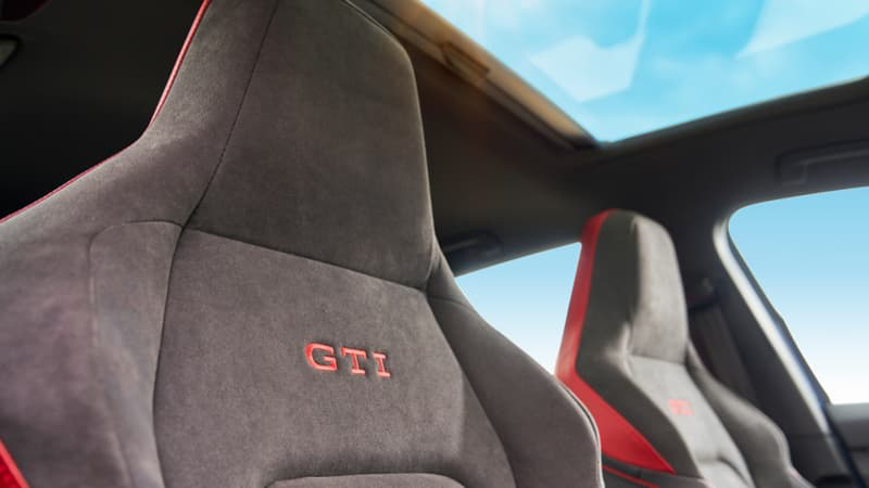 Close-up of available front sport seats in a Volkswagen Golf GTI with GTI logo stitching, gray suede material, and red Kings Red Metallic accents, under an available panoramic sunroof